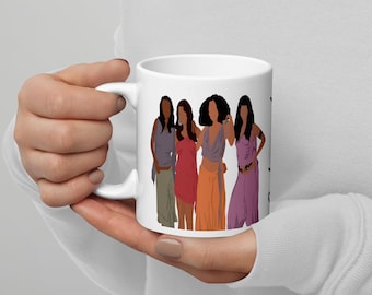May include: White ceramic mug featuring a graphic of four women in various outfits. The women are depicted in shades of brown, red, orange, and purple. The mug is held by a person.
