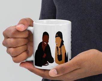 May include: White ceramic mug featuring a minimalist illustration of two people. One person is wearing a black outfit, and the other is wearing overalls and a cap. The mug is held by a person.