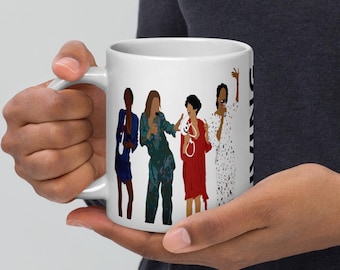 May include: White ceramic mug featuring a graphic of four women in colorful outfits. The women are depicted in a minimalist style, with each wearing a different outfit. The mug has a curved handle and a black text on the side.