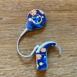 May include: A white hearing aid with a blue and orange fish design. The hearing aid has a small, round microphone and a curved ear hook.