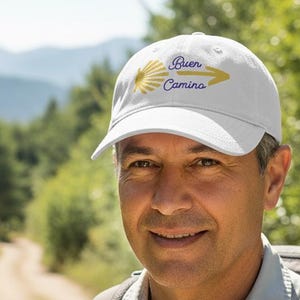 May include: White baseball cap with the text "Buen Camino" in blue and yellow. The cap has a curved peak and is worn by a person on a hiking trail. The background includes a dirt path, trees, and mountains.