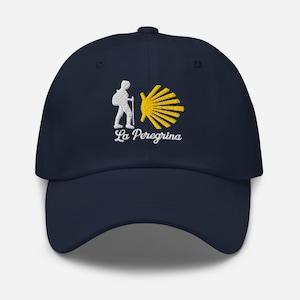 May include: Navy blue baseball cap featuring an embroidered design. The design includes a white pilgrim figure with a walking stick, a yellow scallop shell, and the text "La Peregrina" in white script. The cap has a curved brim.