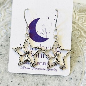 May include: Silver star-shaped earrings with a fairy design. The earrings are hanging from a white card with the text "Wandering Bunny Artisan Handmade Jewelry".