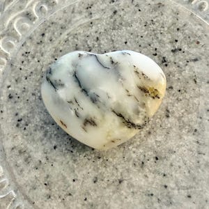 May include: A white heart-shaped stone with black and green veins. The stone is sitting on a gray, textured surface.