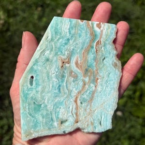 May include: A flat, polished slab of Caribbean calcite featuring a vibrant turquoise and light blue color palette with swirling, organic patterns of tan and white mineral inclusions throughout the stone's surface and small natural cavities.