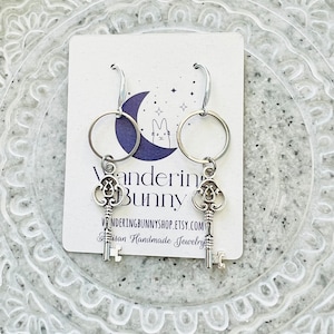May include: Silver-toned key earrings with a circular hoop design. The earrings hang from a white card with the text "Wandering Bunny" and "Artisan Handmade Jewelry". The background is a decorative, light gray plate.