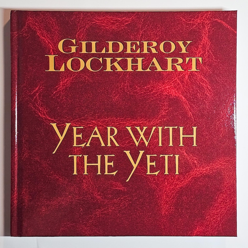 Puede incluir: Un libro de tapa dura cuadrado con una cubierta de color rojo intenso y texturizada con un patr&oacute;n jaspeado. El t&iacute;tulo GILDEROY LOCKHART YEAR WITH THE YETI est&aacute; impreso en letras may&uacute;sculas doradas con serifa en el centro de la portada.