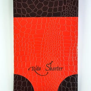 May include: A rectangular item with a faux alligator skin pattern. The top and bottom sections are dark brown, the middle is bright red, and the text "Rita Skeeter" is in the centre. The item has a glossy finish.