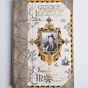 Puede incluir: Un libro de tapa dura titulado "Gilderoy Lockhart Travels with Trolls". La portada presenta un retrato en un marco dorado, un diseño de mapa y detalles en pan de oro. El libro tiene un lomo texturizado y estampado.