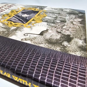 Puede incluir: Un libro de tapa dura titulado Year with the Yeti de Roy Hart. La portada presenta una ilustraci&oacute;n en blanco y negro de una escena forestal con un marco dorado ornamentado que contiene un retrato, y un lomo texturizado morado oscuro.