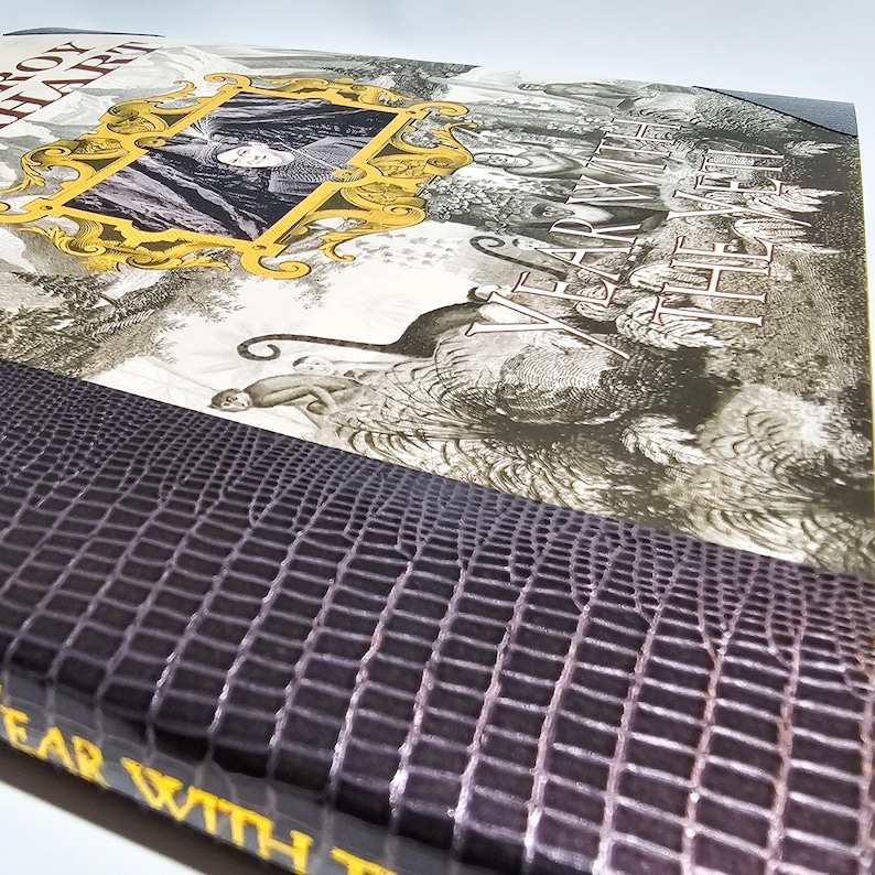 Puede incluir: Un libro de tapa dura titulado Year with the Yeti de Roy Hart. La portada presenta una ilustraci&oacute;n en blanco y negro de una escena forestal con un marco dorado ornamentado que contiene un retrato, y un lomo texturizado morado oscuro.