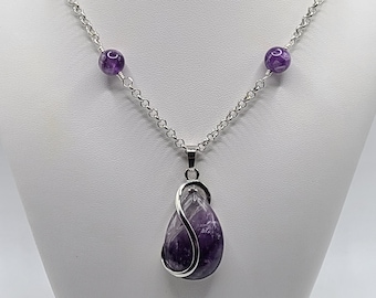 May include: A silver chain necklace with a large purple amethyst pendant. The pendant is wrapped in silver wire and has a teardrop shape. The necklace is adorned with smaller purple amethyst beads.