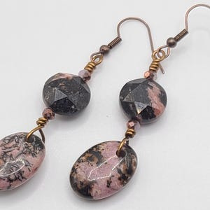 May include: A pair of dangle earrings featuring faceted black and pink gemstone beads. The earrings have oval-shaped pink and black stones at the bottom, connected by copper-colored wire and hooks. The earrings are designed for pierced ears.