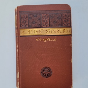 May include: A vintage book titled "Indian Summer" by W.D. Howells. The cover is a deep reddish-brown with gold lettering and decorative floral patterns. The book has a worn appearance, suggesting age and use.