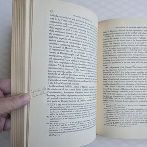 May include: Open book with text on aged paper. The pages are yellowed, and the text is in black. A hand is holding the book open, revealing the text and the book's binding. The book appears to be a historical document.