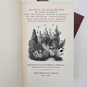 May include: Open book with the title "Journal of Researches" by Charles Darwin. The cover features an illustration of marine life and text. Published by The Heritage Press, New York.