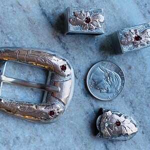 May include: Silver-toned belt buckle and matching accessories with a floral design and red gemstone accents. The set includes a buckle, two rectangular pieces, and a small decorative piece. A U.S. dime is included for size reference.