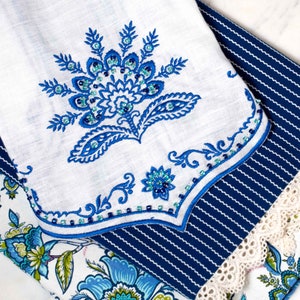 May include: A white tea towel with a blue and green floral embroidery design. The towel is folded and layered on top of a blue and white striped fabric with a white lace trim.