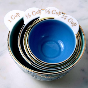 May include: A set of four blue and white ceramic measuring cups with gold trim. The cups are labeled 1 cup, 1/2 cup, 1/3 cup, and 1/4 cup.