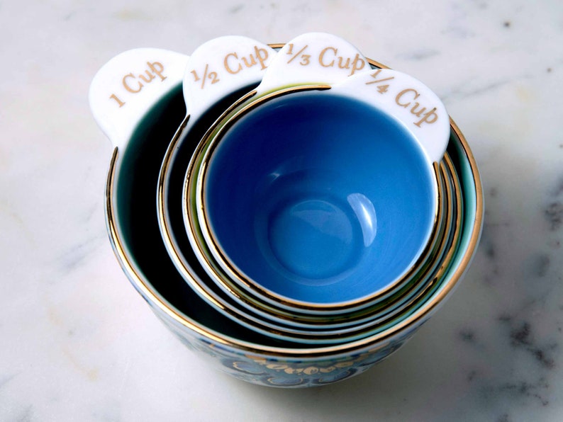 May include: A set of four blue and white ceramic measuring cups with gold trim. The cups are labeled 1 cup, 1/2 cup, 1/3 cup, and 1/4 cup.