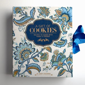 May include: A white cookbook with a blue and gold floral design. The title is "A Gift of Cookies: Recipes to Share with Family & Friends" by Mary Disomma.