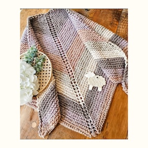 May include: A hand-knitted triangular shawl in shades of pink, brown, and beige with a decorative stitch pattern. The shawl is lying on a wooden surface with a small white ceramic sheep figurine.