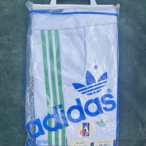 May include: A pair of white Adidas shorts with three green vertical stripes, packaged in a clear plastic bag with a white hanger. The shorts feature a blue Adidas trefoil logo and text, an ATP logo, and a label reading Milano M6 8648 size 34/85.
