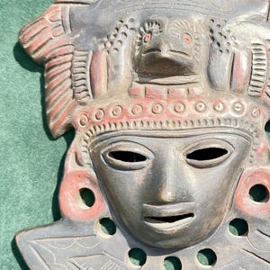 May include: A dark gray and reddish-brown clay mask featuring a central face with almond-shaped eyes and a mouth opening. The forehead displays a bird figure, and the lower section has circular cutouts and etched line patterns.