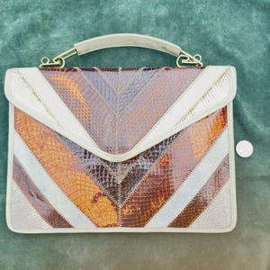 May include: A rectangular handbag featuring a chevron pattern made of alternating panels of brown, tan, and cream-colored snakeskin-textured material. The bag has a top handle with gold-tone metal hardware and a flap closure. A small coin is visible.