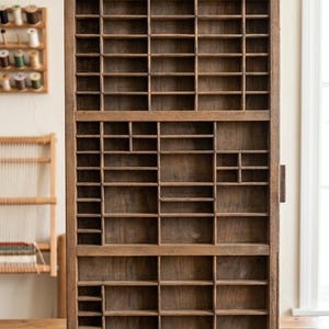 May include: A vintage wooden printer's tray with numerous small compartments. The tray is made of aged wood, showcasing a natural, weathered appearance. It features a rectangular shape with a handle on the side, ideal for organizing small items or displaying collectibles.