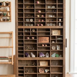 May include: A vintage wooden type tray filled with antique keys, buttons, dried flowers, crystals, and found objects. The tray has small compartments and labels that read "Vintage Type Tray", "Found Objects", and "Wotitory Shierds". The sign above reads "Shop Heritage Finds Co."