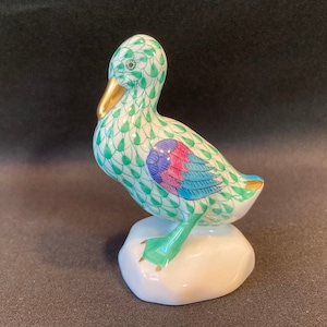 May include: A green and white porcelain duck figurine with blue and pink accents. The duck is standing on a white base.