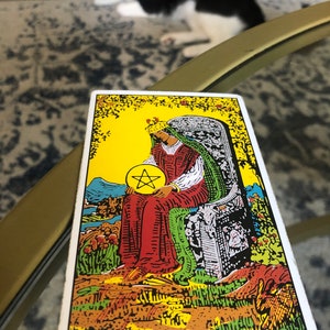 May include: Queen of Pentacles tarot card featuring a woman wearing a crown and a green robe sitting on a throne holding a gold pentagram. The card has a yellow background with green and red accents.