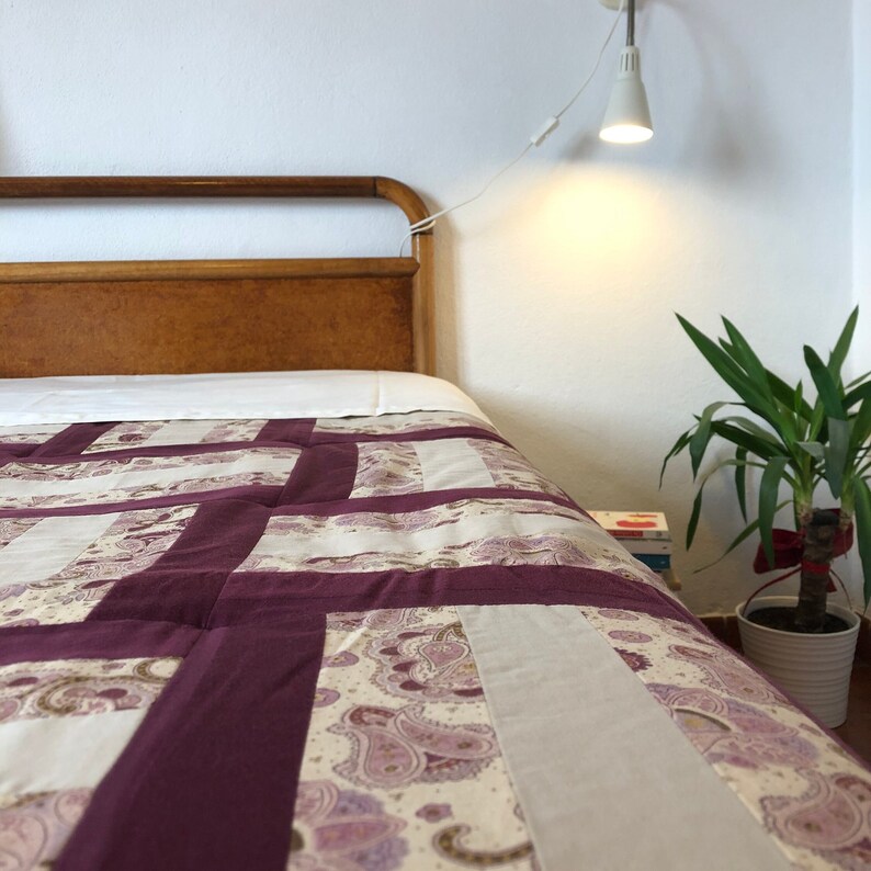 May include: A bed with a wooden headboard and a purple and white patchwork quilt. The bed is made up with white sheets and a white pillow. A lamp is on the wall behind the bed.