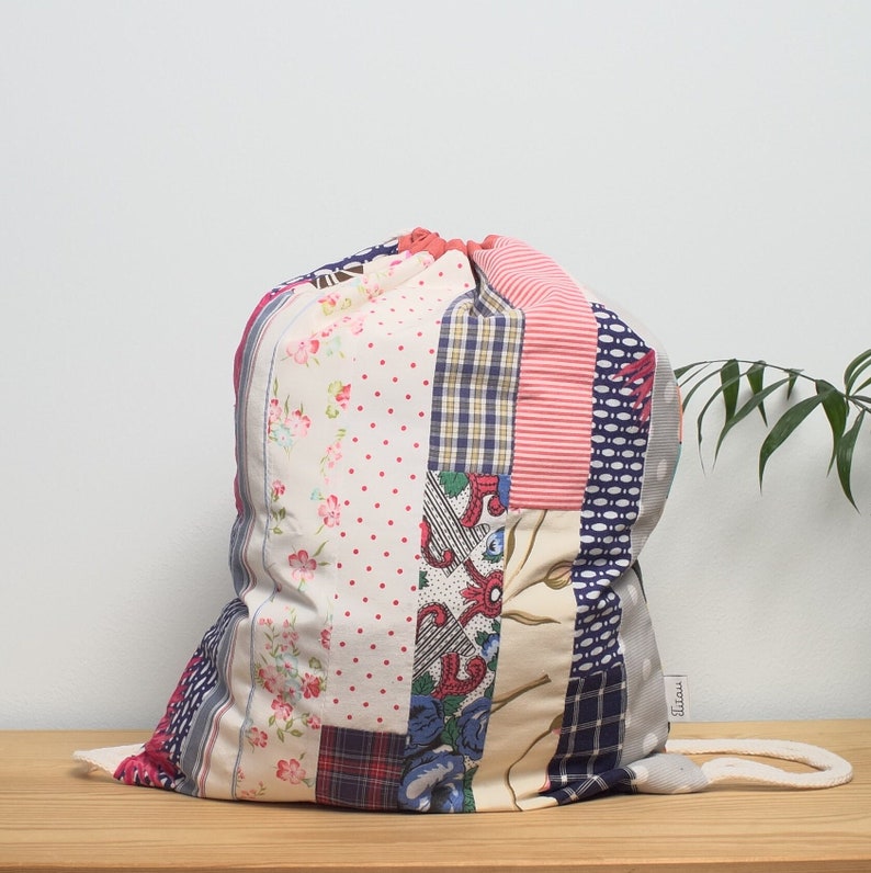 K&ouml;nnte beinhalten: Eine Patchwork-Kordeltasche aus verschiedenen Stoffen in Rosa-, Blau- und Wei&szlig;t&ouml;nen. Die Tasche hat ein Blumenmuster, Punkte und Karomuster. Die Tasche hat einen wei&szlig;en Kordelzug und ein wei&szlig;es Etikett mit dem aufgedruckten Wort "Tilda".