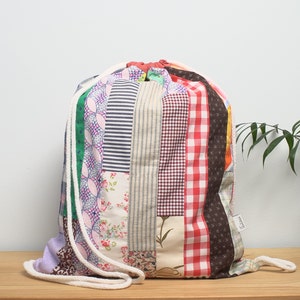 Puede incluir: Una bolsa de cordón de retazos con una variedad de telas coloridas, incluyendo rojo, blanco, azul, verde y marrón. La bolsa tiene un cordón blanco y una pequeña etiqueta que dice "Elara".