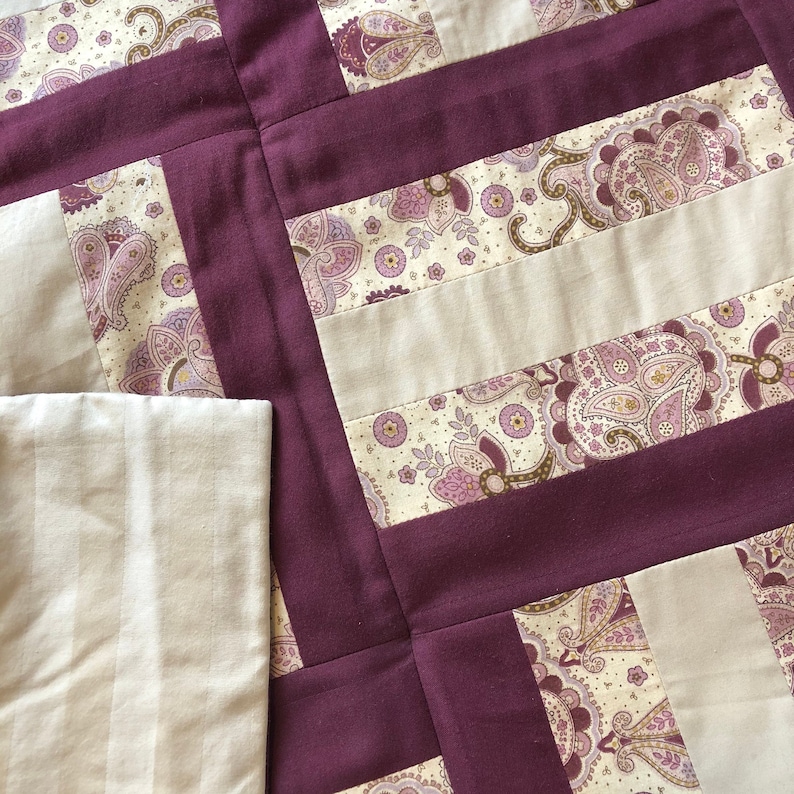 May include: A quilt with a patchwork design featuring a burgundy background and a cream-colored paisley pattern. The quilt is made up of squares and rectangles, and the seams are visible.