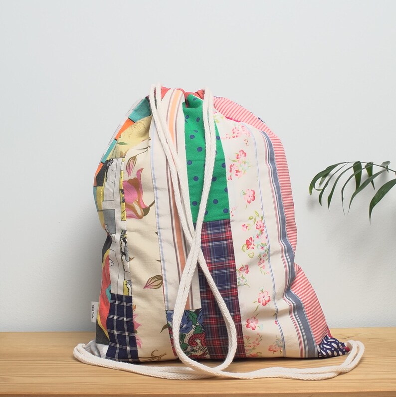 K&ouml;nnte beinhalten: Eine bunte Patchwork-Kordeltasche mit einem wei&szlig;en Baumwollkordel. Die Tasche ist aus verschiedenen Stoffen mit Blumen-, Streifen- und Karomustern gefertigt.