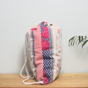 K&ouml;nnte beinhalten: Eine wei&szlig;e Kordeltasche mit rosa und blauen Blumen- und Streifenmustern. Die Tasche hat einen wei&szlig;en Kordelverschluss und ist aus weichem Baumwollstoff gefertigt.