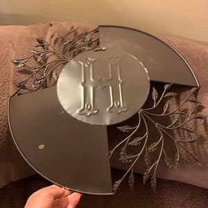 May include: A metal wall decor featuring a large letter "H" surrounded by a wreath of stylized leaves. The wreath is made of thin metal and has a dark brown finish.