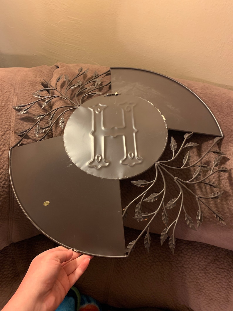 May include: A metal wall decor featuring a large letter "H" surrounded by a wreath of stylized leaves. The wreath is made of thin metal and has a dark brown finish.