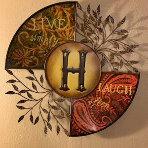 May include: A metal wall decor with a circular design featuring the letter "H" in the center. The design is divided into three sections, each with a different pattern and text. The top section is green with the text "Live simply". The right section is orange with the text "Laugh often". The left section is brown with a floral pattern.