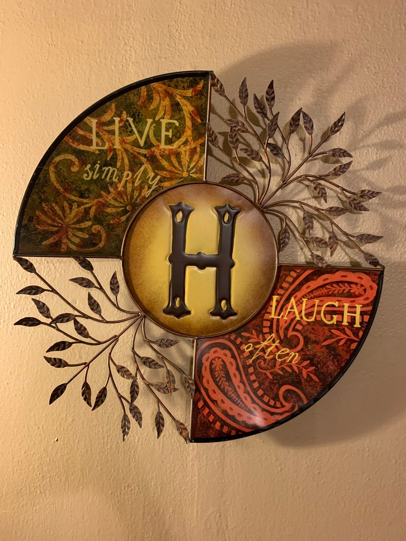May include: A metal wall decor with a circular design featuring the letter "H" in the center. The design is divided into three sections, each with a different pattern and text. The top section is green with the text "Live simply". The right section is orange with the text "Laugh often". The left section is brown with a floral pattern.