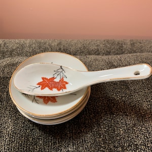 Könnte beinhalten: Ein Set aus vier weißen Suppenschalen aus Keramik mit goldfarbenem Rand und einem passenden weißen Suppenlöffel aus Keramik mit einem roten Ahornblatt-Design.