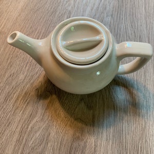 May include: A small, cream-coloured ceramic teapot with a matching lid. The teapot has a curved spout, a rounded body, and a handle. The lid features a curved handle. The teapot is designed for brewing tea.