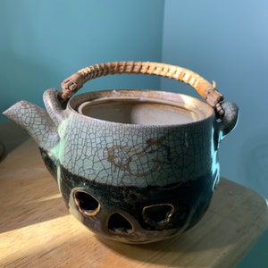 Może przedstawiać: Ceramiczny czajnik z pękniętą glazurą i plecionym bambusowym uchwytem. Czajnik ma ciemnobrązową podstawę i jasnoszary wierzch z złotym wzorem. Czajnik ma trzy otwory na spodzie.