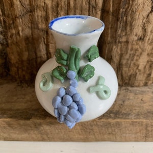 May include: A small white ceramic vase with a blue rim and a 3D design of grapes and leaves. The grapes are a light purple color and the leaves are green.