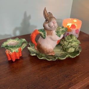 Puede incluir: Un juego de servir de cerámica con temática de Pascua. Cuenta con una tetera con forma de conejito con asas de zanahoria y una base con forma de lechuga. También hay una cremaera a juego con forma de zanahoria y una fuente cubierta. El juego está decorado con colores verdes, marrones, naranjas y blancos.