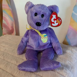 May include: Purple Beanie Baby bear with a yellow ribbon and an Easter egg design. The bear has black eyes and a black nose. The tag reads "Ty Beanie Babies".
