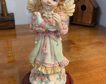 Może przedstawiać: Porcelanowa figurka anioła z blond włosami i różową sukienką trzymająca niebieskiego ptaka. Anioł stoi na chmurze z różowymi różami.